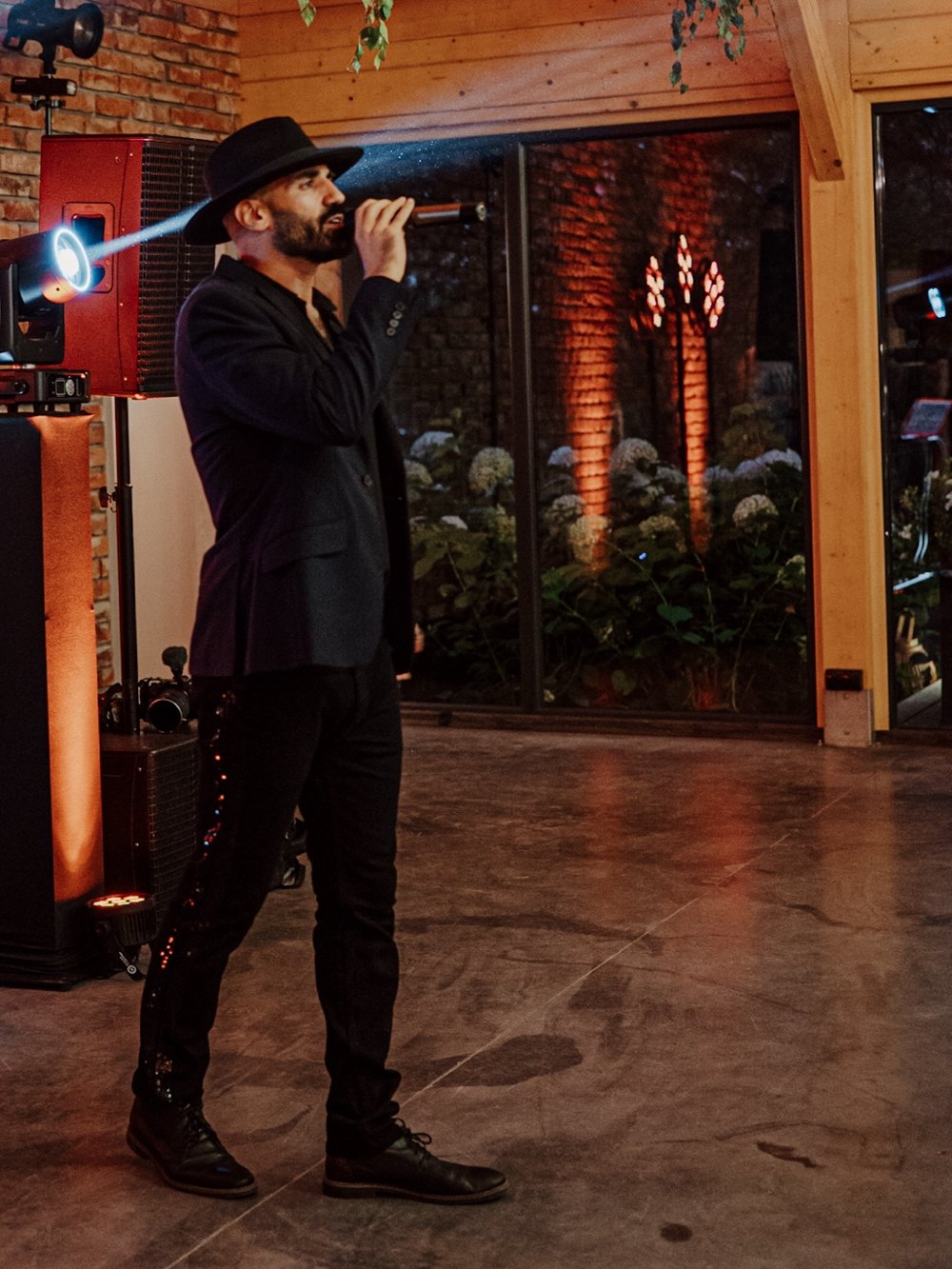 Sami Harb in a black fedora singing under stage lights at an evening event